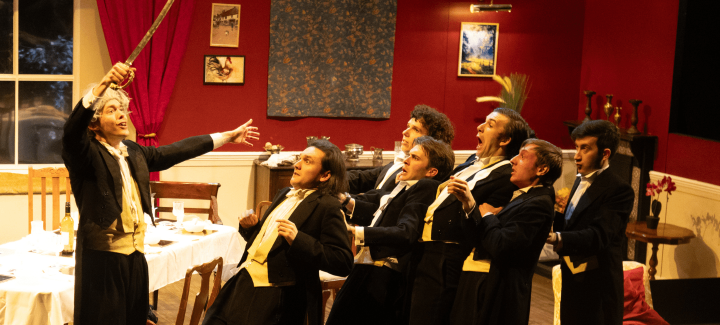 A group of men in suits recoil from an older man wielding a sword.
